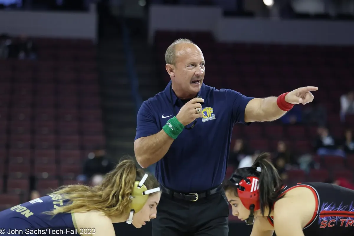 An official in a navy polo points as two wrestlers set up in a neutral stance.