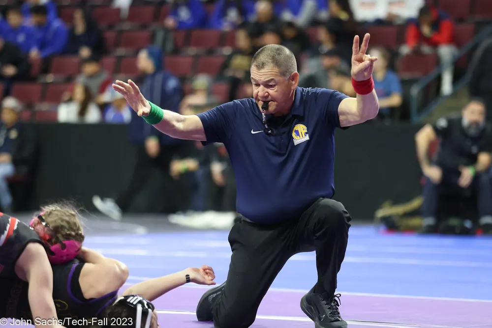 An official kneels at the edge of the mat and signals near-fall points.